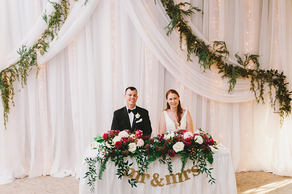 wedding sweatheart table with white  backdrop, greenery and lush flowers now & always