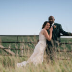 Casual boho wedding, bride and groom hugging looking at the camera, they are leaning on a stone wall